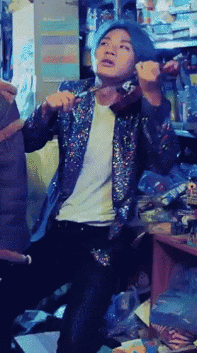 a man wearing a sequined jacket and a blue hat is standing in a cluttered room
