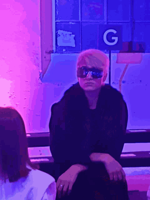 a woman in sunglasses sits in front of a sign that says g