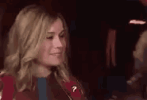 a woman is sitting in a dark room and smiling while holding her hand to her chin .