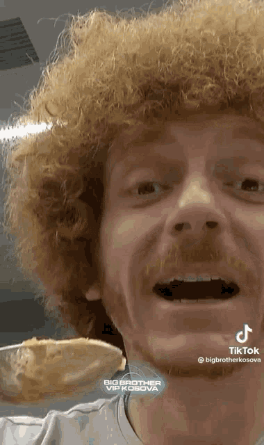 a man with curly hair and a beard is holding a piece of bread in front of his mouth .
