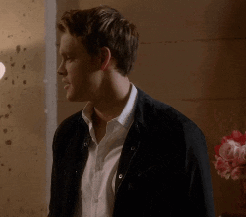 a man wearing a black jacket and a white shirt stands in front of a vase of flowers
