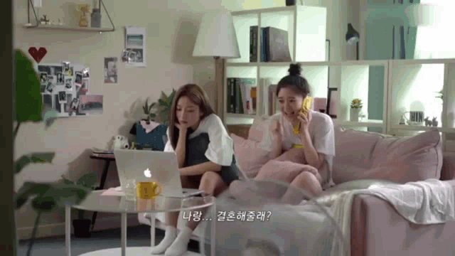 two women are sitting on a couch looking at a laptop and one is talking on a cell phone