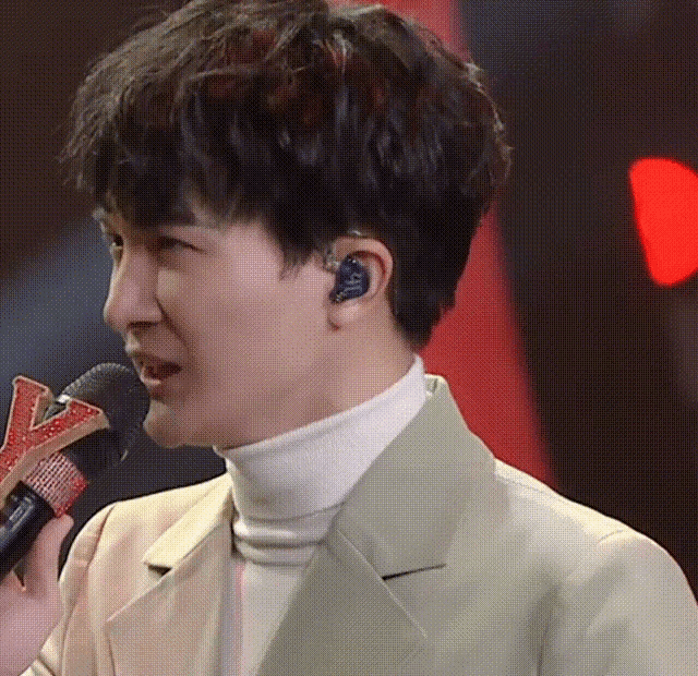 a man wearing a white turtleneck and a tan jacket is singing into a microphone .