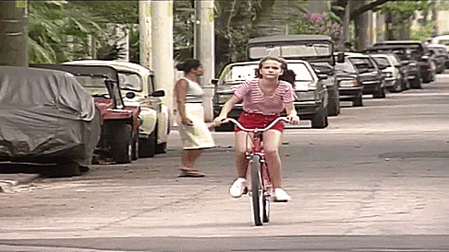a girl is riding a bike down a street with cars parked behind her