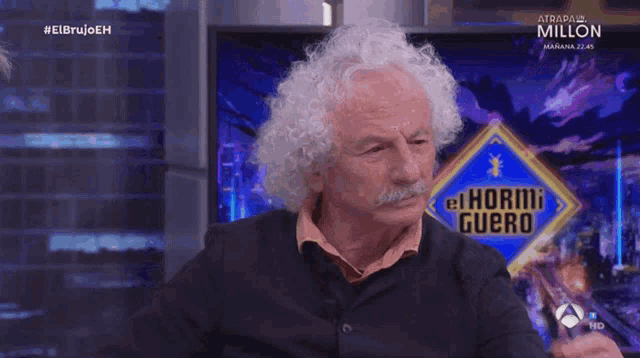 a man with curly hair and a mustache is sitting in front of a tv screen that says #elbrujoeh