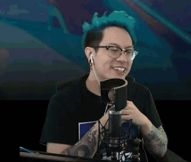 a man with blue hair and glasses is sitting in front of a microphone with his fist in the air .
