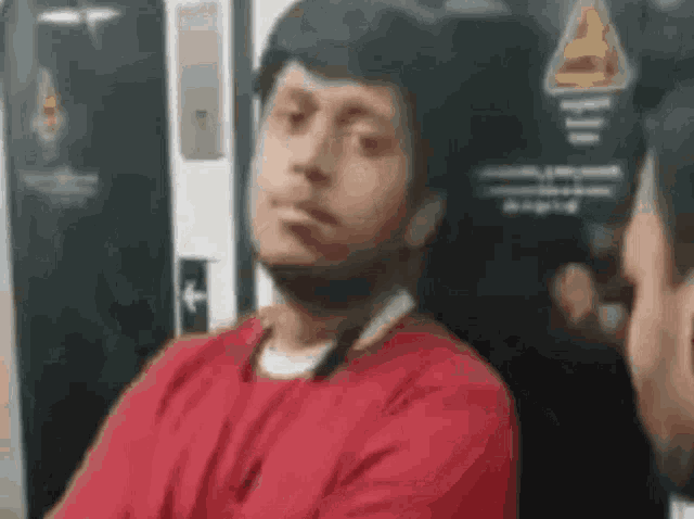 a man in a red shirt is sitting on a train with his arms crossed and looking at another man .