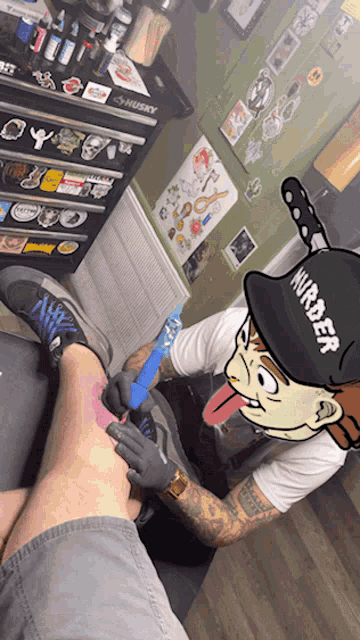 a man is getting a tattoo on someone 's leg and the man is wearing a hat that says murder