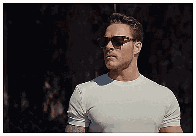 a man wearing sunglasses and a white shirt stands in front of a fence