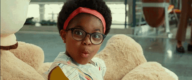 a little girl wearing glasses and a red headband is sitting next to a stuffed animal