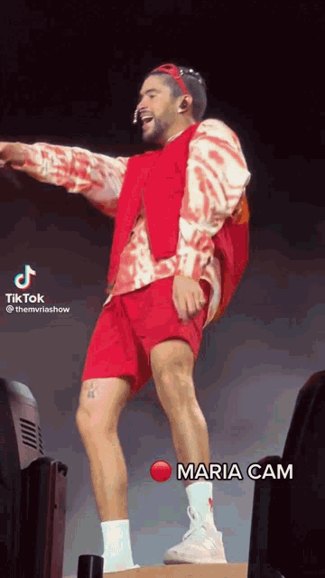 a man in a red shirt and shorts is dancing on a stage with a red button that says maria cam