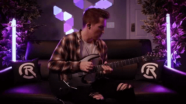 a man is sitting on a couch playing a guitar with purple lights behind him .