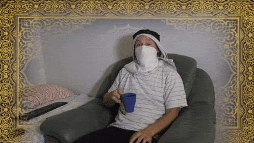 a man with a scarf around his face sits in a chair holding a cup