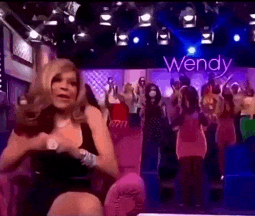 a woman in a black dress is dancing in front of a crowd with the word wendy on the wall behind her .