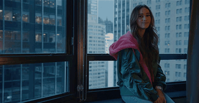 a woman in a green jacket sits on a window sill in front of a window