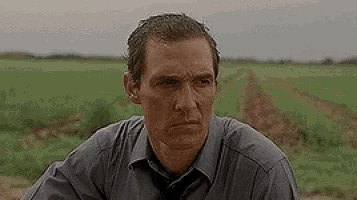 a man in a gray shirt and tie is sitting in a field looking at the camera .