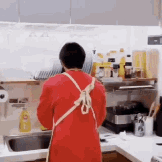 a man wearing a red apron is standing in a kitchen washing dishes .