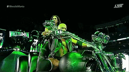 a wrestler is riding a green motorcycle in front of a sign that says wrestlemania live