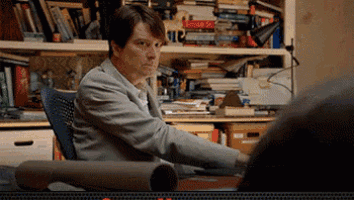 a man in a suit sits at a desk in front of a bookshelf full of books