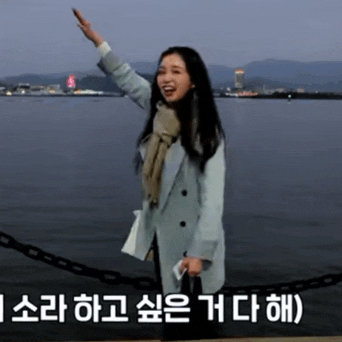 a woman in a blue coat is standing in front of a body of water and waving