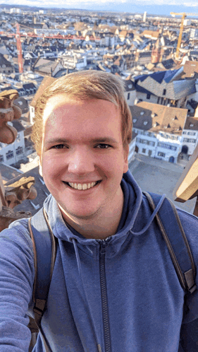a young man in a blue hoodie smiles in front of a cityscape