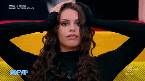 a woman in a black turtleneck holds her hair in front of a yellow and red flag with #gfvip written on the bottom