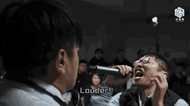a man holding a microphone says louder in front of a crowd of people