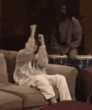 a man is playing drums while a woman sits on a couch with her arms in the air