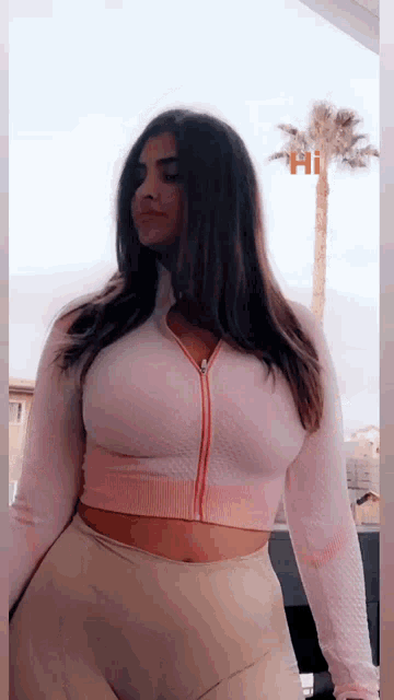 a woman is standing in front of a palm tree wearing a pink crop top and tan pants .