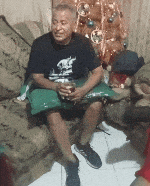 a man is sitting on a couch holding a glass and a pillow with a christmas tree in the background