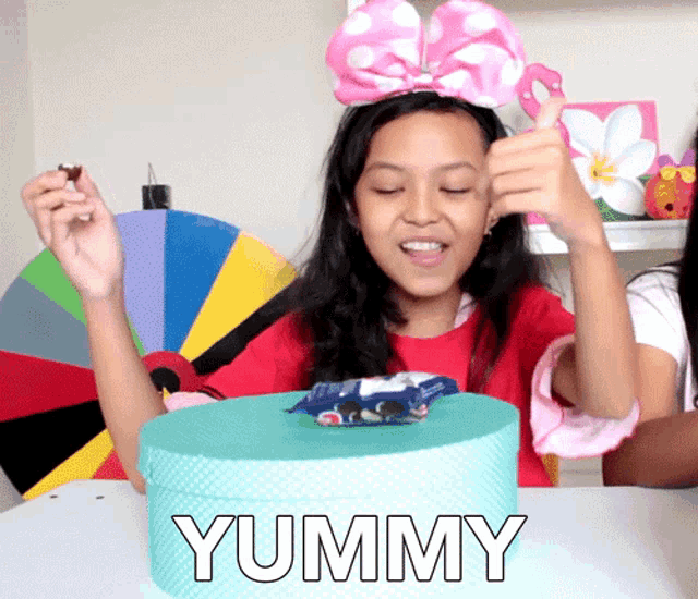 a girl with a pink bow on her head is sitting at a table with a box that says yummy in front of her