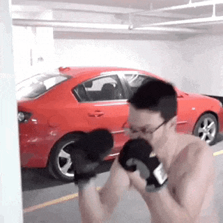 a shirtless man is wearing boxing gloves in front of a red car in a garage .