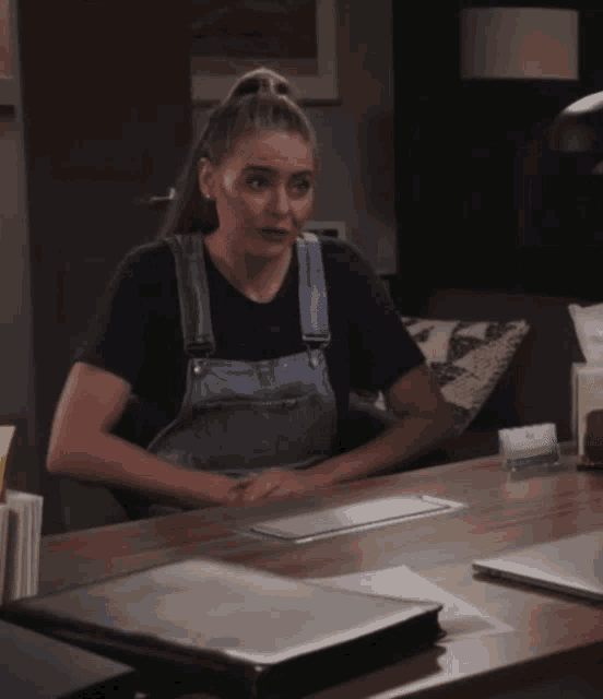 a woman wearing overalls sits at a desk with a laptop