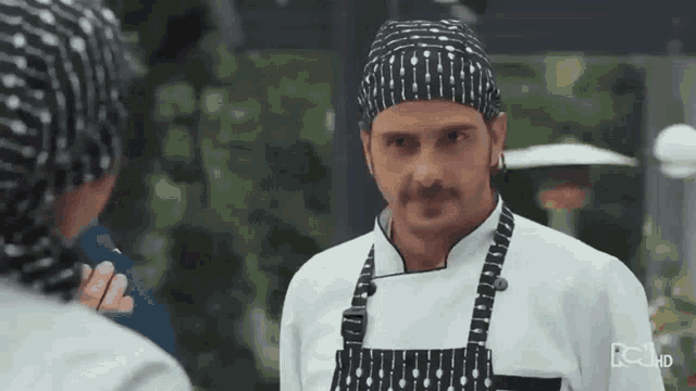 a man in a chef 's uniform and apron is talking to another man .