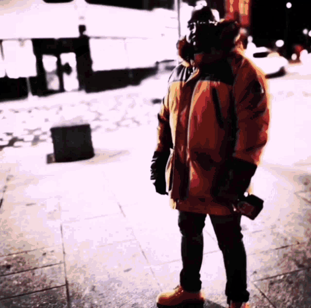a man in an orange parka is standing on a sidewalk holding a bottle
