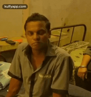 a man in a grey shirt is sitting on a bed in a room .