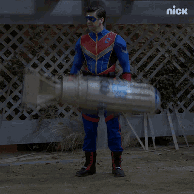 a man in a superhero costume is holding a large object in front of a fence with the nick logo on the bottom