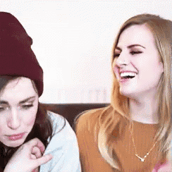 two women are sitting next to each other on a couch and laughing . one of the women is wearing a beanie .