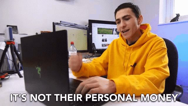 a man in a yellow hoodie is typing on a laptop with the words " it 's not their personal money " above him