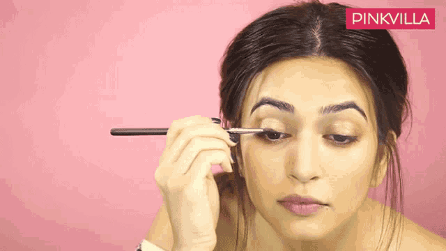 a woman is applying eyeliner with a pinkvilla logo in the background
