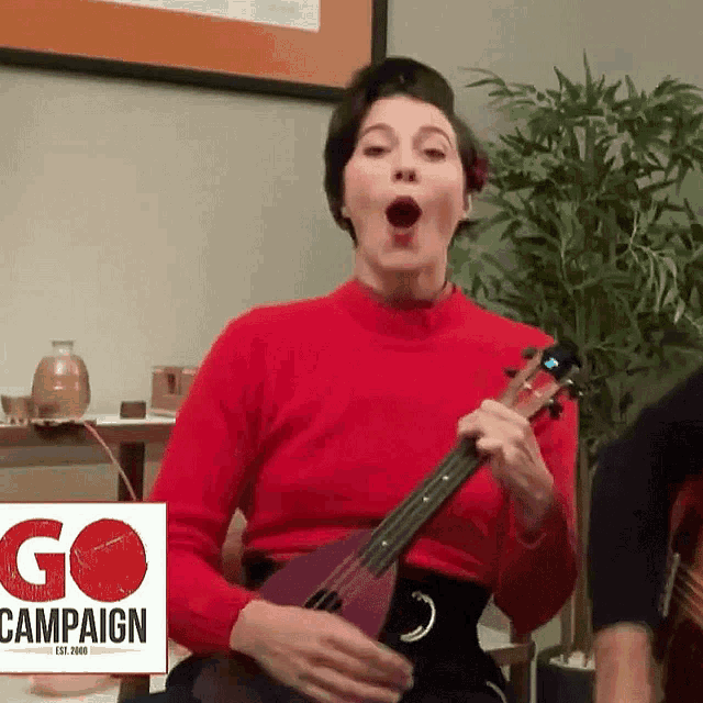 a woman in a red sweater is playing a ukulele in front of a go campaign sign