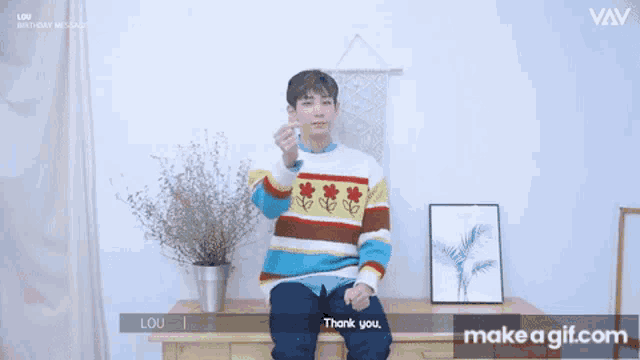 a man in a colorful sweater is sitting at a table and giving a heart sign .