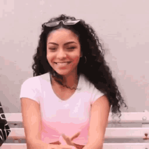 a woman with curly hair is sitting on a bench wearing sunglasses and a pink shirt .