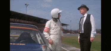 a man in a suit and hat talks to a man in a race suit