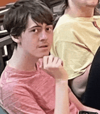 a young man in a pink shirt is sitting in a chair with his hand on his chin and looking at the camera .