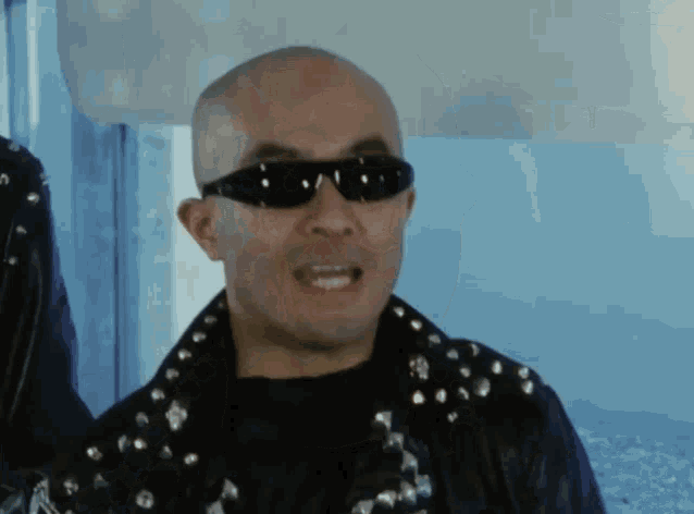 a bald man wearing sunglasses and a black jacket with studs