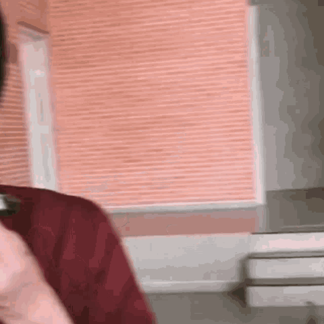 a man in a red shirt is holding a cell phone in front of a wall with blinds .