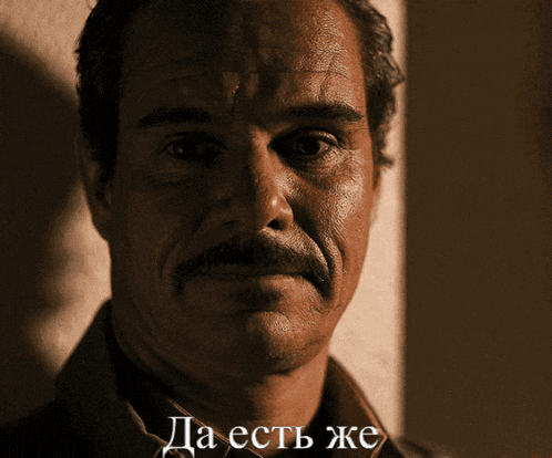 a man with a mustache is smiling in a dark room with the words да есть же written below him