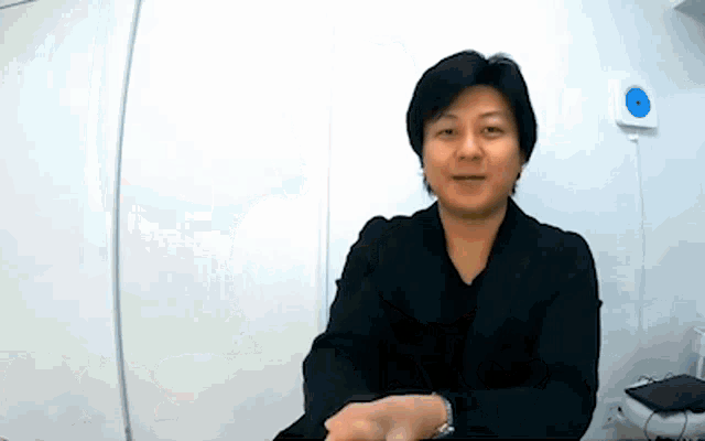a man in a black jacket is sitting in front of a white wall and smiling