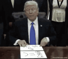 a man in a suit and tie is sitting at a table holding a piece of paper that says " donald trump "